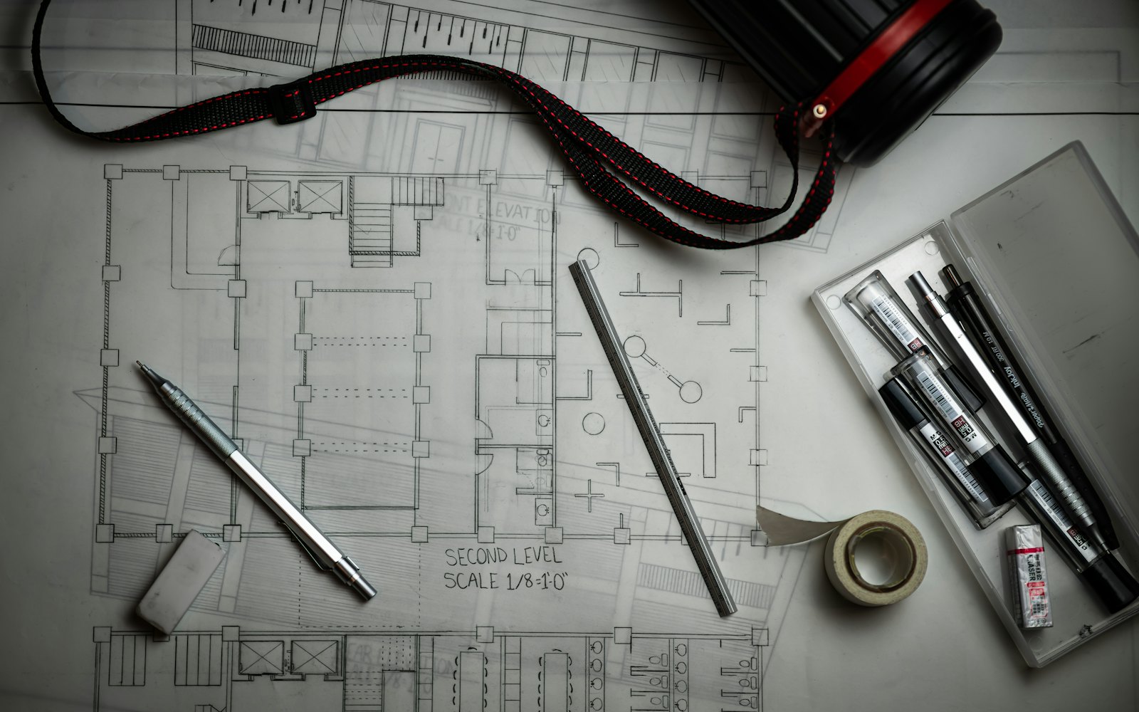 A pencil-drawn floor plan on a drafting table with a mechanical pencil, a scale ruler, tape and pens